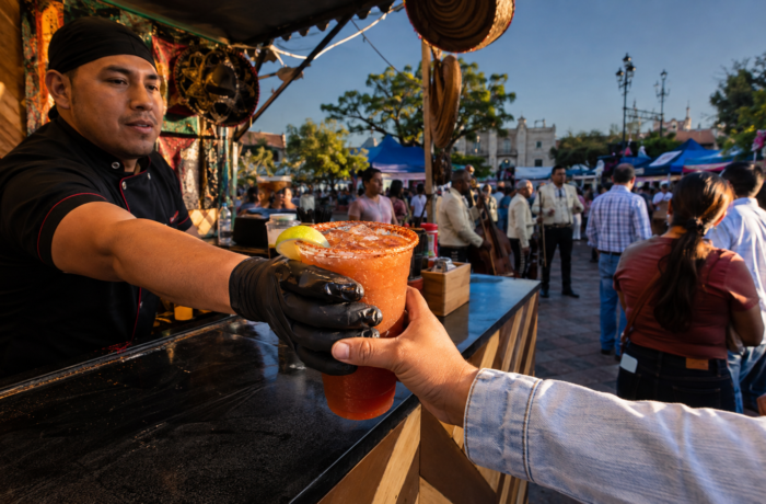Una michelada escarchada pasa de una barra a manos de una persona mientras músicos y puestos de comida se preparan en una plaza de Villagrán